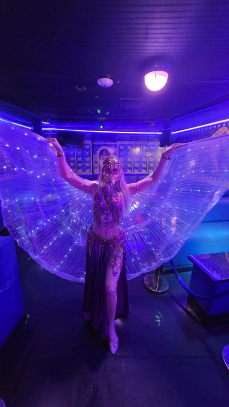 A masked dancer wearing illuminated wings in a purple and blue lounge